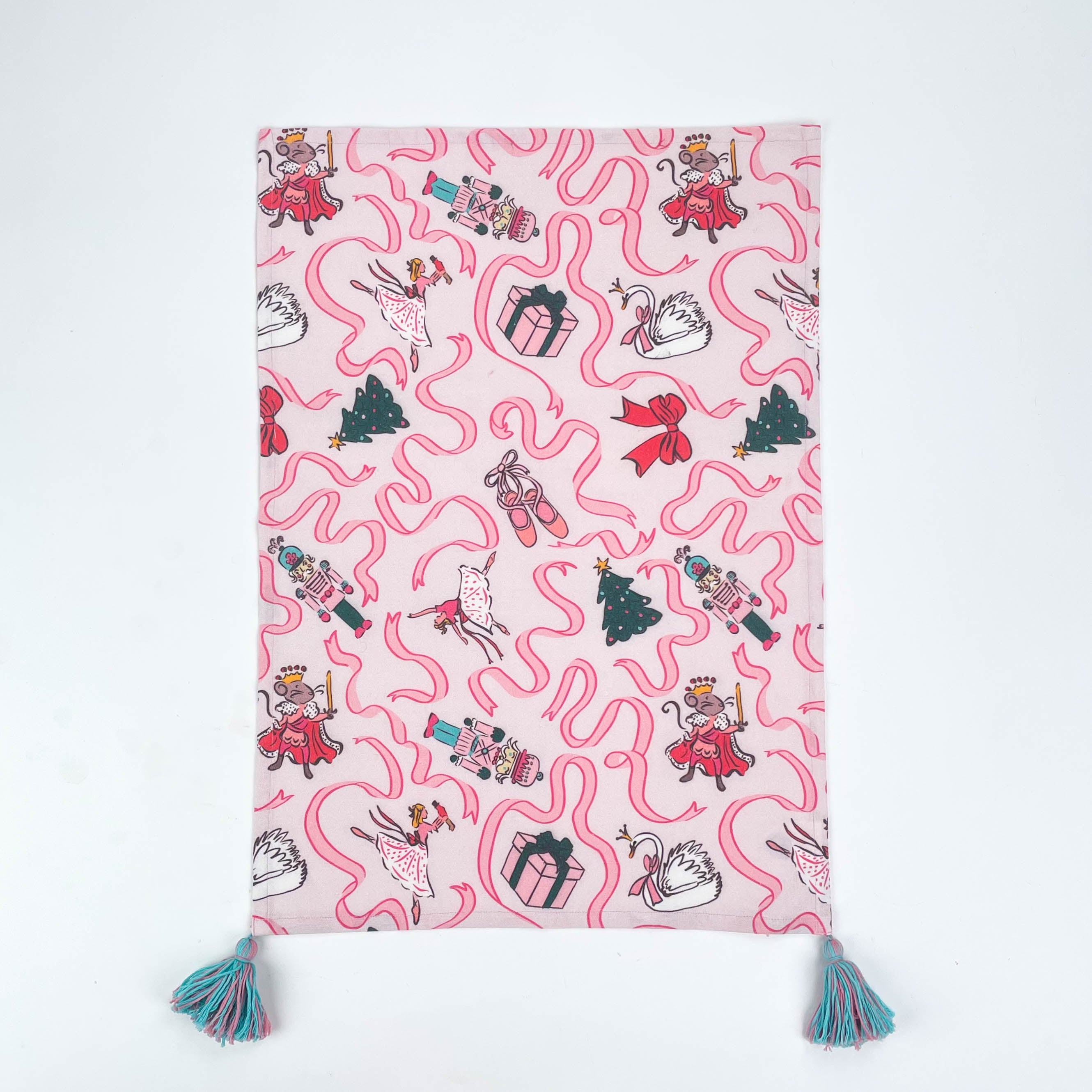 Pink Christmas tea towel featuring Nutcracker-themed illustrations with ribbons, ballerinas, swans, Christmas trees, and presents, finished with teal tassels on each corner.
