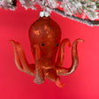 An orange glass octopus christmas ornament hanging against a red background