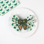 Green and white patterned cloth napkin folded into a bow with a woven brown ribbon napkin ring displayed on a white plate.