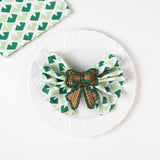 Green and white patterned cloth napkin folded into a bow with a woven brown ribbon napkin ring displayed on a white plate.