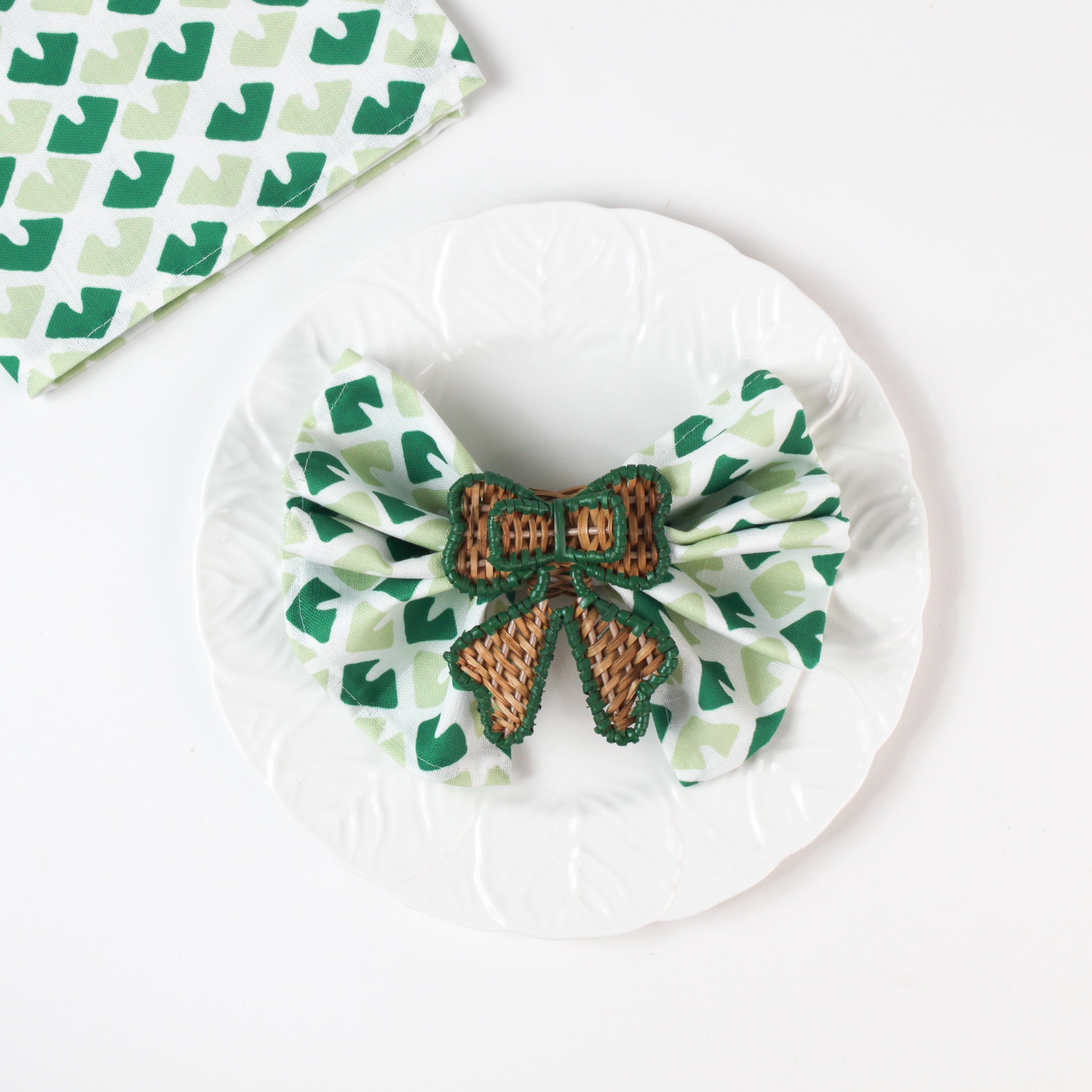 Green and white patterned cloth napkin folded into a bow with a woven brown ribbon napkin ring displayed on a white plate.