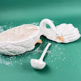 White ceramic swan-shaped serving tureen with detachable ladle, textured feather detailing, shown on green backdrop with snow accents.
