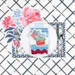 Patriotic table setting featuring a hydrangea and American flag place card, floral placemat, white dinner plate, and blue patterned napkin.