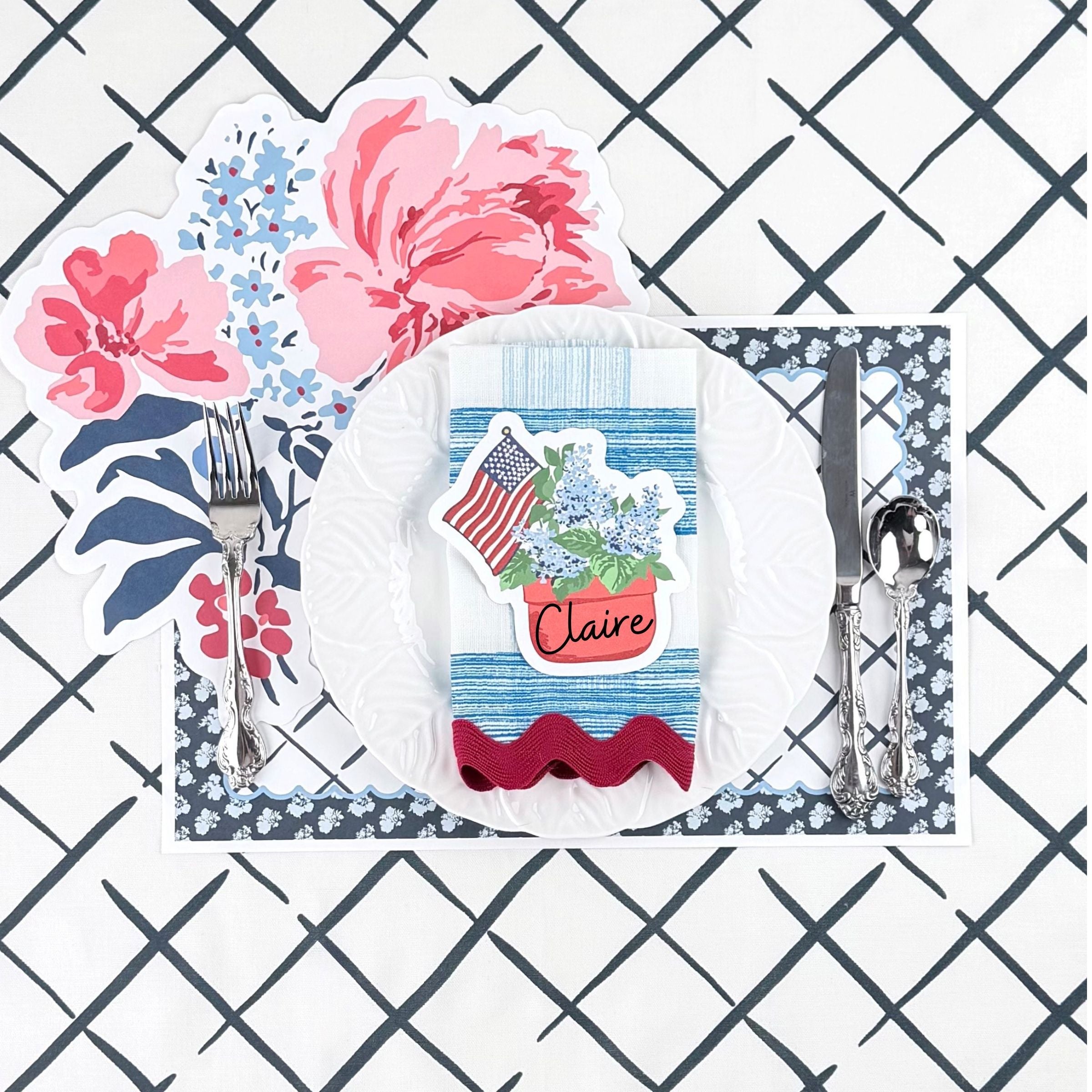 Patriotic table setting featuring a hydrangea and American flag place card, floral placemat, white dinner plate, and blue patterned napkin.