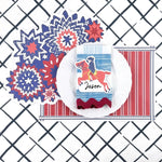 A styled patriotic table setting featuring a red white and blue starburst paper placemat, striped placemat, white plate, decorative napkin, and place card on a navy trellis patterned background.