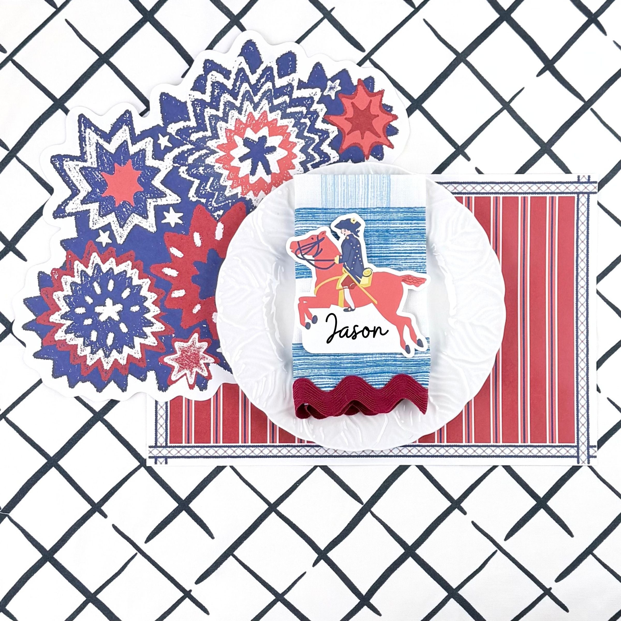 A styled patriotic table setting featuring a red white and blue starburst paper placemat, striped placemat, white plate, decorative napkin, and place card on a navy trellis patterned background.