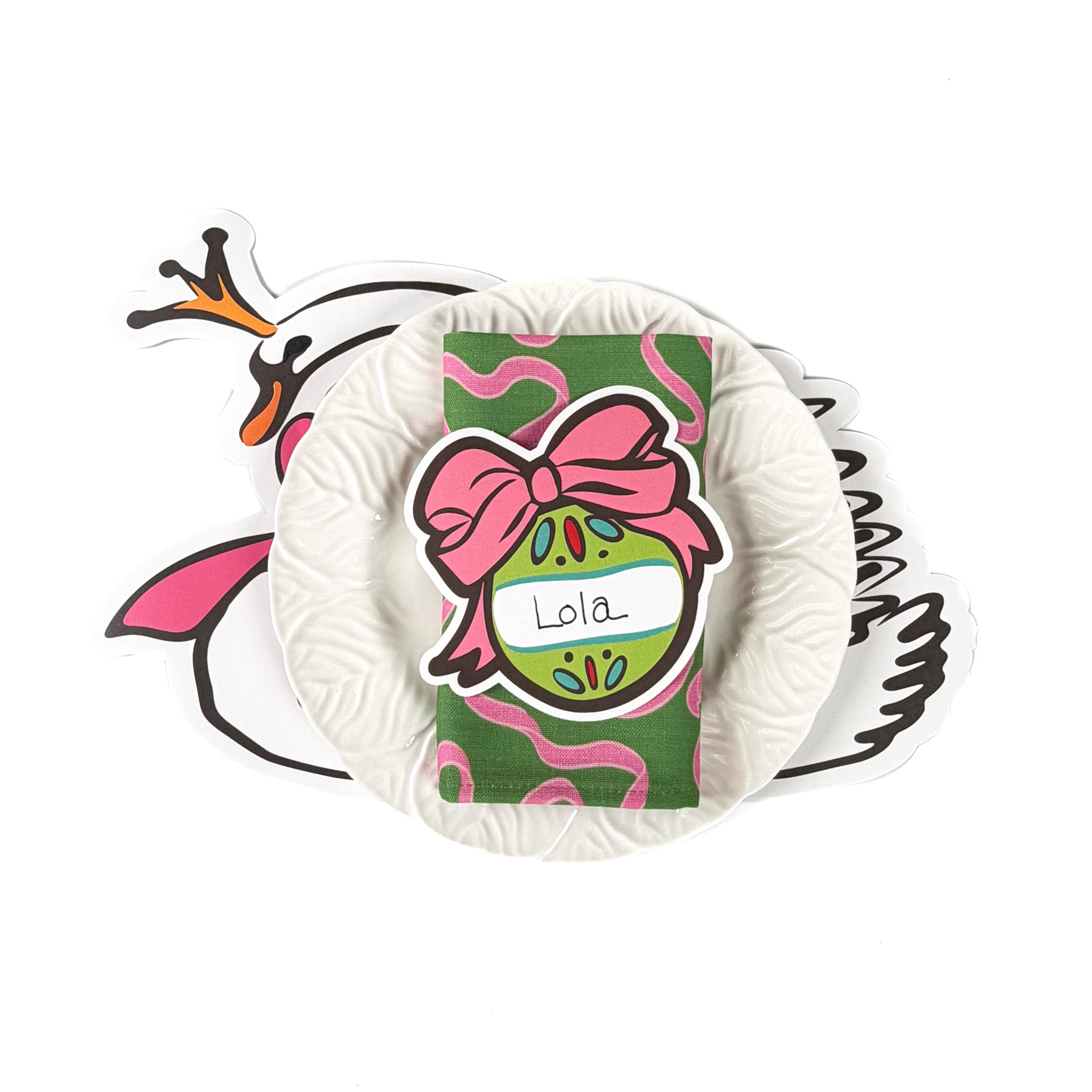 Christmas tabletop display featuring pink bow ornament–shaped paper napkins, a white ruffled plate, and a green holiday napkin with personalized ornament design, arranged on a white background.