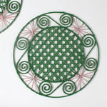 a pink and green round rattan placemat with green swirls and pink star woven motifs against a white backround.