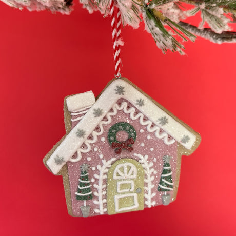 a small pink gingerbread house ornament with white detailing and christmas trees hanging against a red background