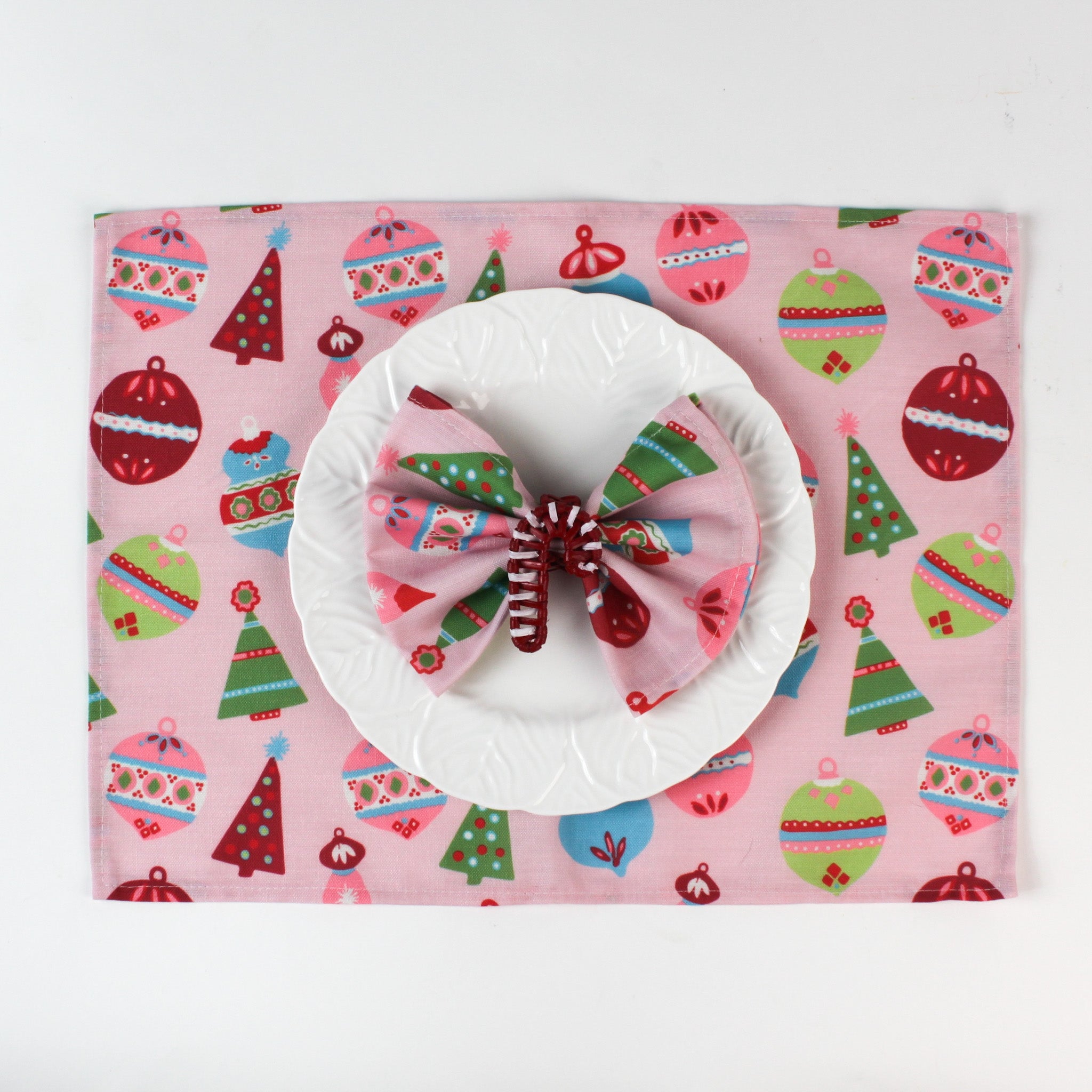 a red candy cane christmas napkin ring filled with a pink christmas napkin with white plate and ornament napkin against a white background. 