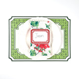 Holiday tabletop place setting featuring a red bordered paper place card with handwritten name, white ruffled plate, floral holiday napkin, and green patterned placemat, displayed on a white background.