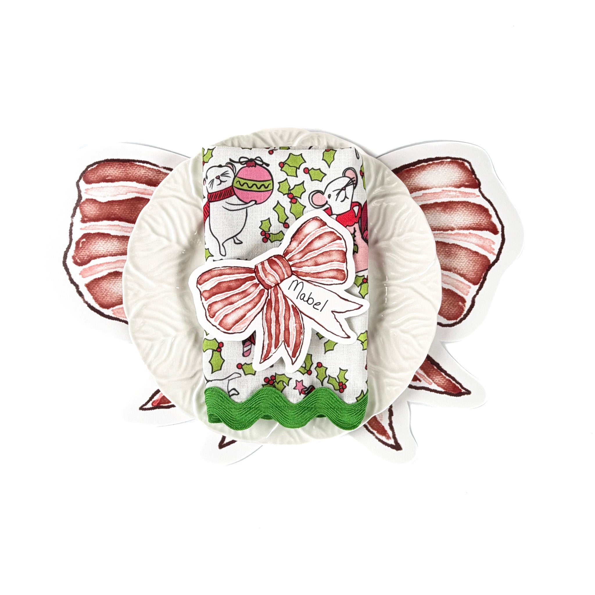 Christmas tabletop display featuring red bow–shaped placecard, a white ruffled plate, and a festive holiday napkin with mouse and holly pattern, arranged on a white background.