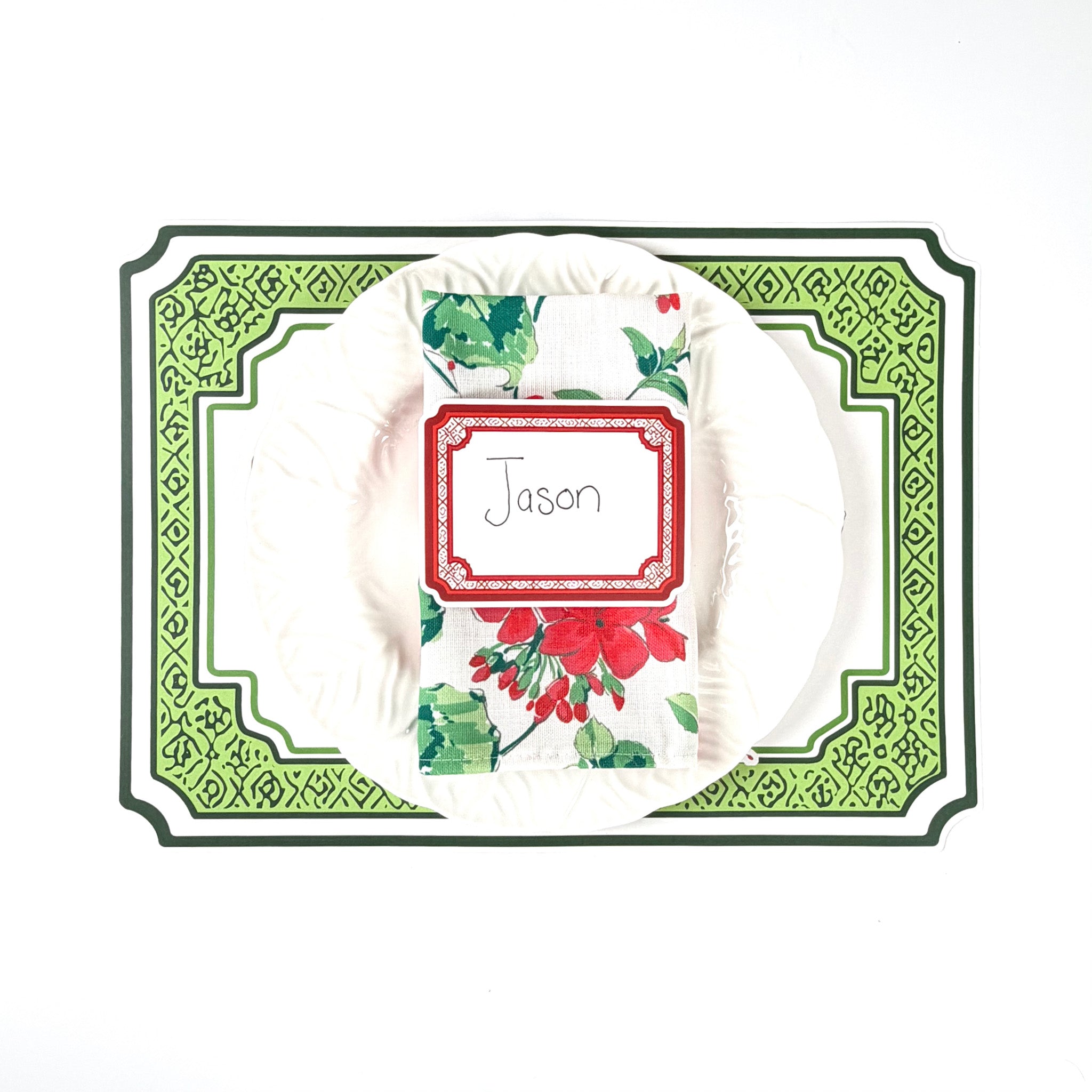 Holiday table setting featuring a red geranium decorative placemat, white ruffled plate, floral holiday napkin, and red bordered paper place card with handwritten name, arranged on a white background.