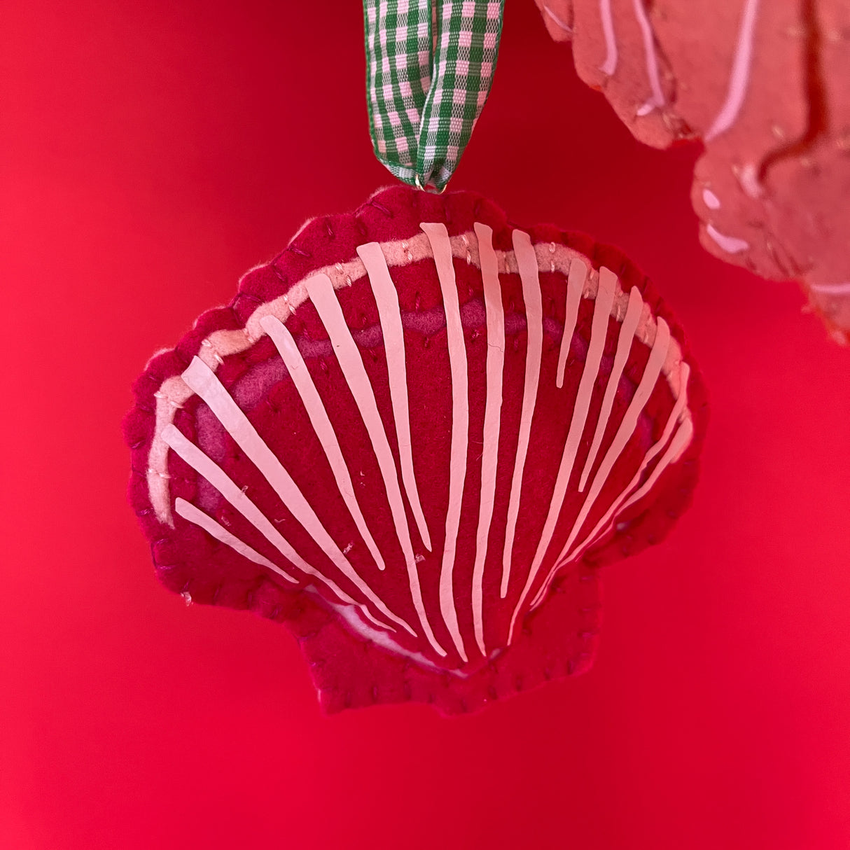 Red felt christmas ornament in the shape of a scallop shell with a green gingham ribbon against a red background