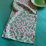 Pink ribbons swirl around on a green background in Madcap Cottage's Ribbons Pink and Green cloth table runner