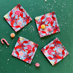 A set of four square cloth cocktail napkins with a red rock lobster wearing a blue bow on a green background, accompanied by candy and confetti.