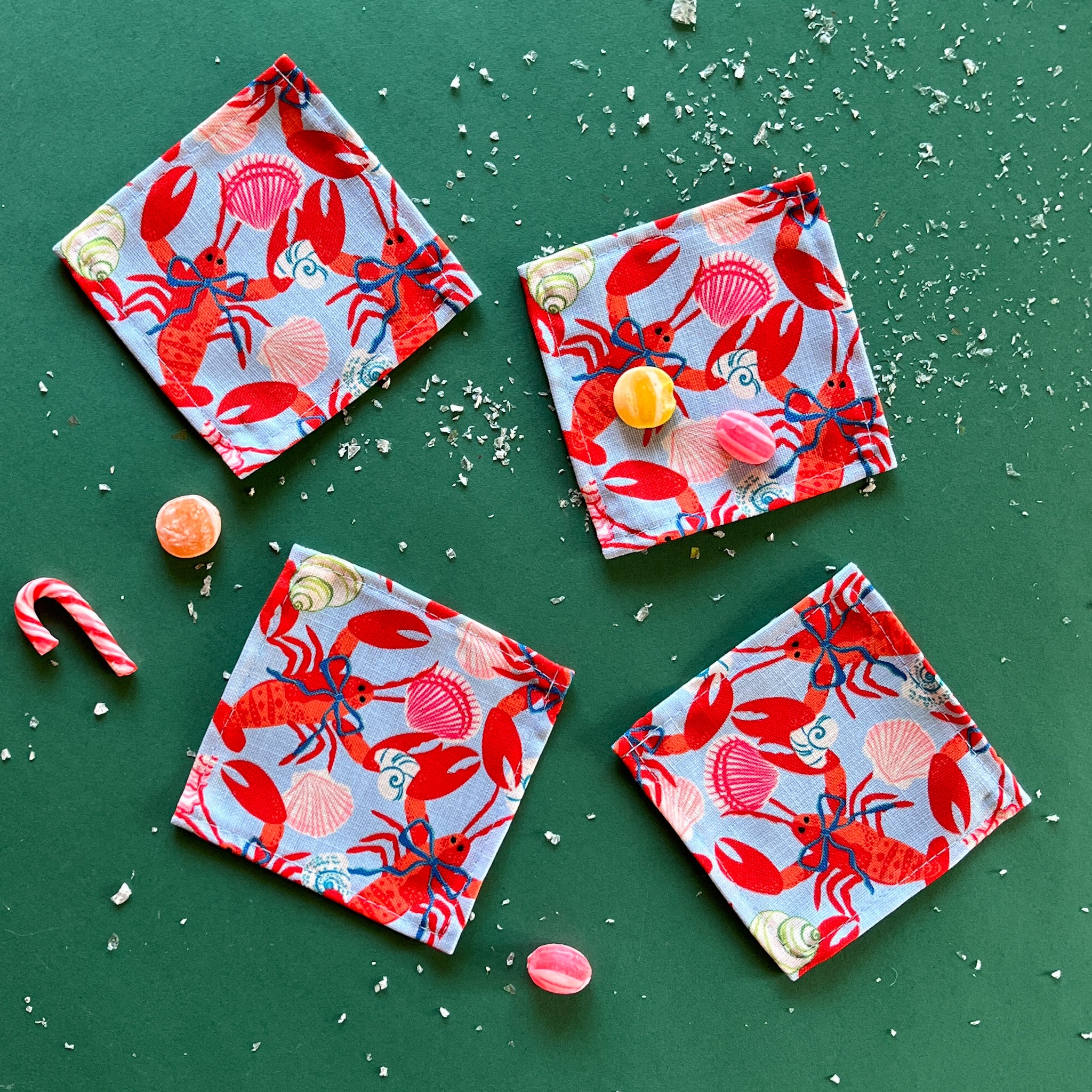 A set of four square cloth cocktail napkins with a red rock lobster wearing a blue bow on a green background, accompanied by candy and confetti.