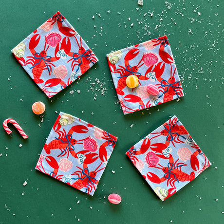 A set of four square cloth cocktail napkins with a red rock lobster wearing a blue bow on a green background, accompanied by candy and confetti.