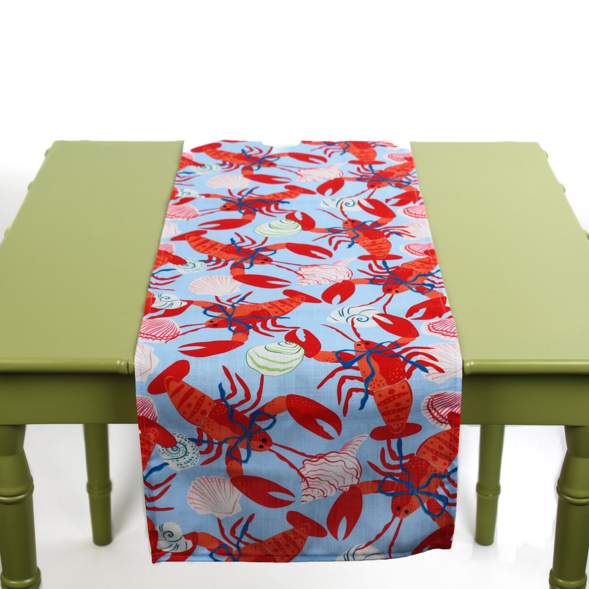 Blue table runner featuring a red lobster and seashell pattern displayed on a green table.