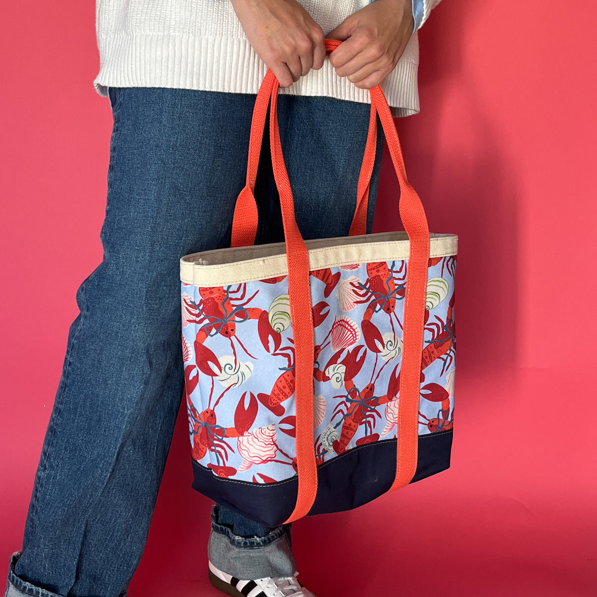 Rock Lobster canvas tote bag with red straps and blue fabric with a repeating lobster and shell motif being held by model
