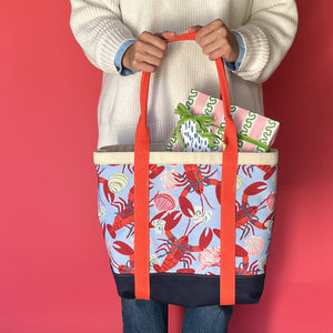 Rock Lobster large canvas tote bag with red straps and blue fabric with a repeating lobster and shell motif being held by model with a present sticking out of tote.