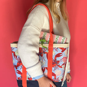 Rock Lobster canvas tote bag with red straps and blue fabric with a repeating lobster and shell motif on models shoulder