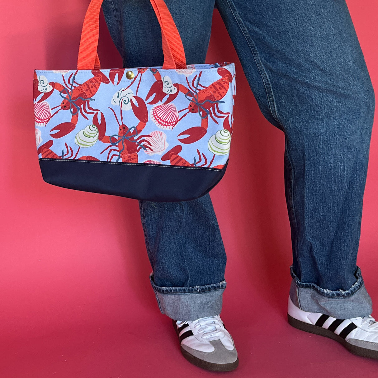 Rock Lobster small canvas tote bag with red straps and blue fabric with a repeating lobster and shell motif hanging by models legs