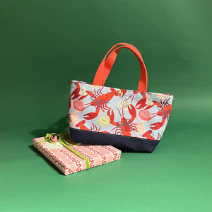 Rock Lobster small canvas tote bag with red straps and blue fabric with a repeating lobster and shell motif resting on a present.