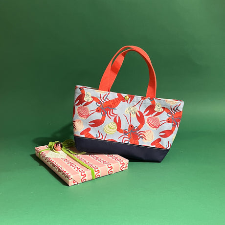 Rock Lobster small canvas tote bag with red straps and blue fabric with a repeating lobster and shell motif resting on a present.