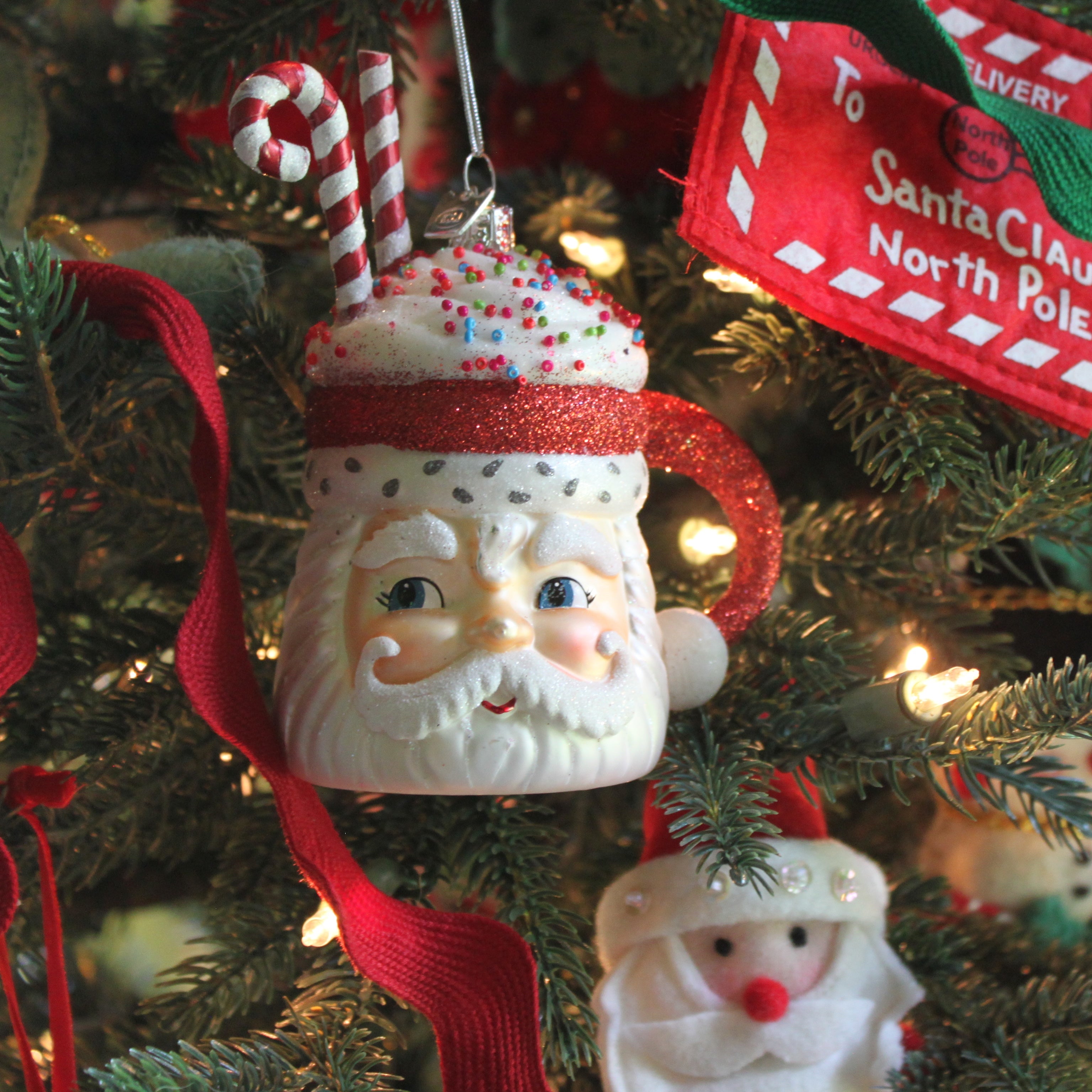 Close-up of a whimsical Santa mug ornament hanging on a Christmas tree, decorated with candy canes, whipped cream details, and colorful sprinkles, surrounded by ribbon and holiday lights.