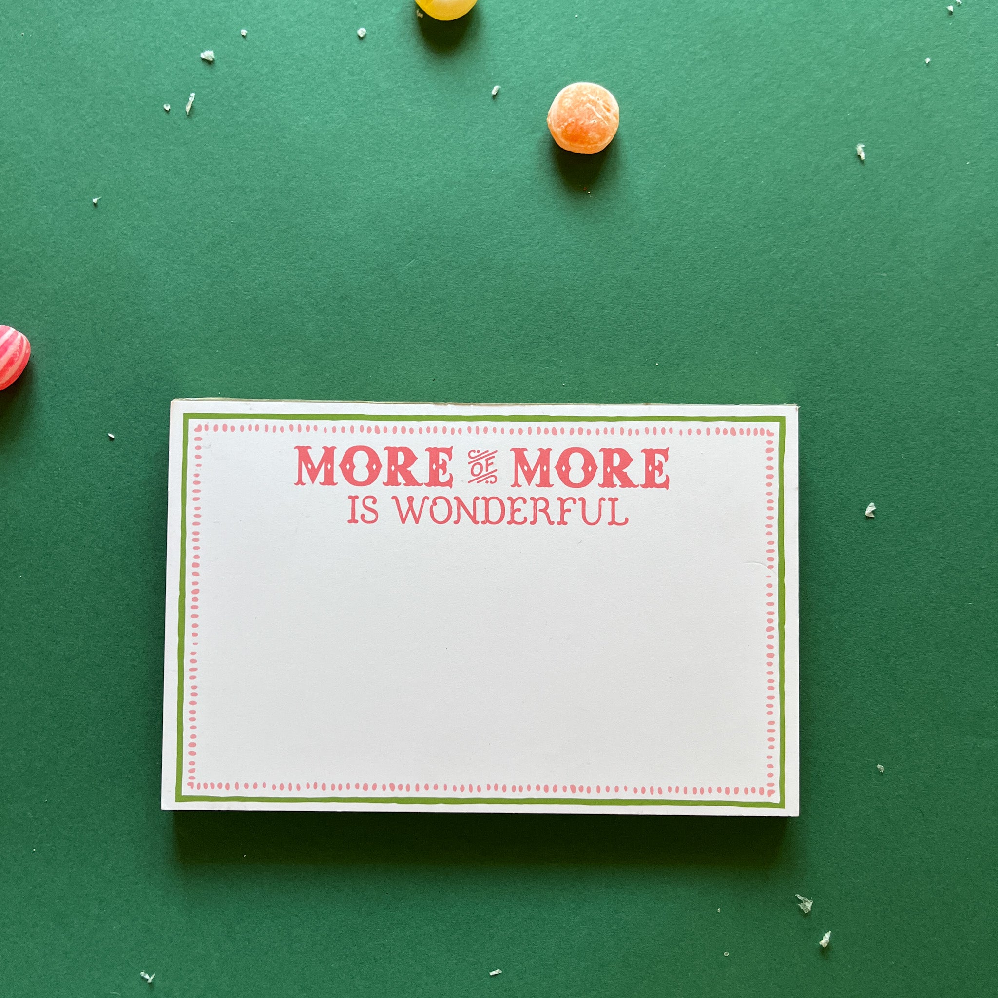 Luxe large notepad with quote ‘More of More is Wonderful’ in red lettering, featuring a dotted border and green trim, perfect for stylish note-taking. placed in a green background