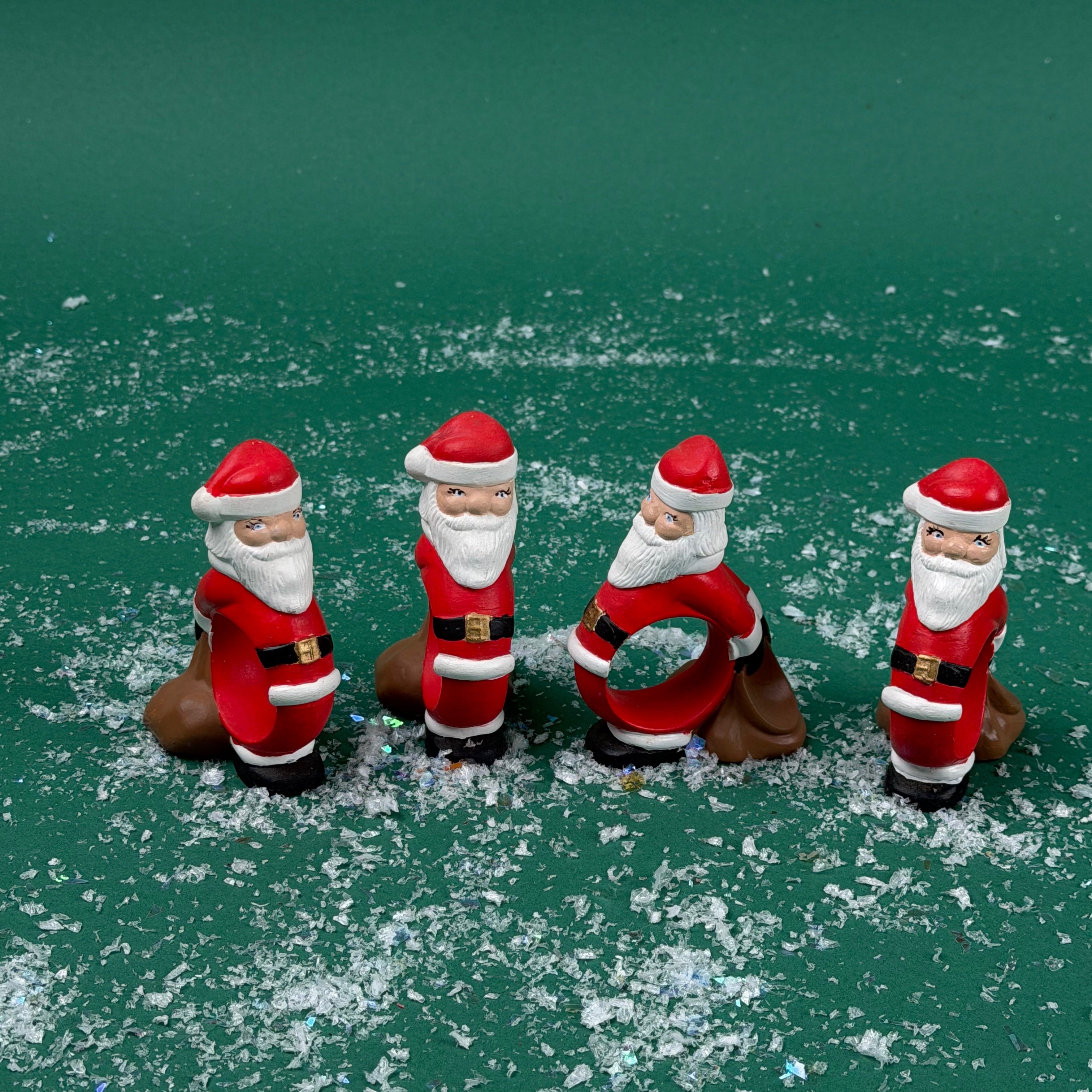Four Santa Claus-shaped napkin rings standing on a green surface with faux snow scattered around