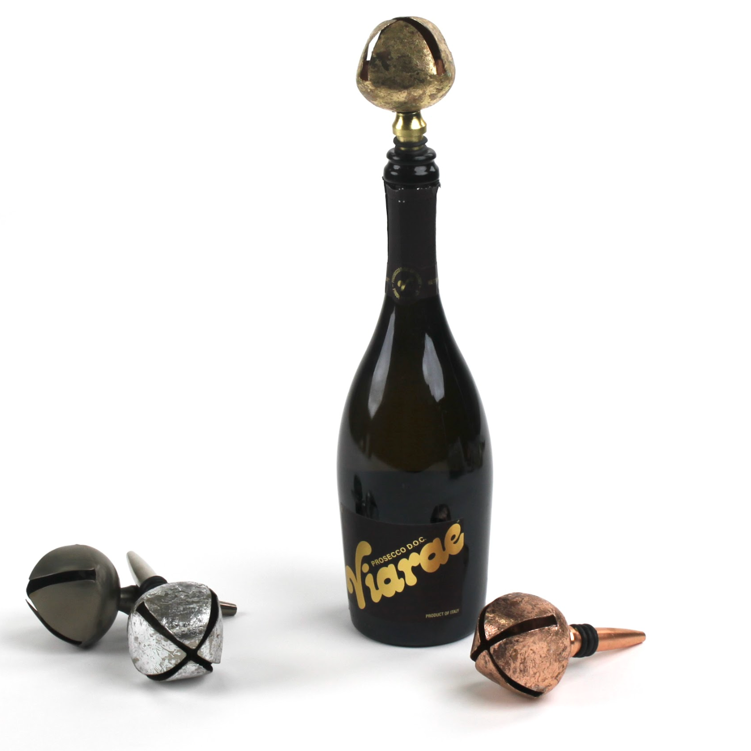four sleighbell christmas bottle stoppers in different metals. a gold one is in a champagne bottle and the three others are resting on a white background. 