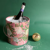 A Shell Seeker ice bucket covered in a pink and green fabric with shell motif and filled with ice and a champagne bottle