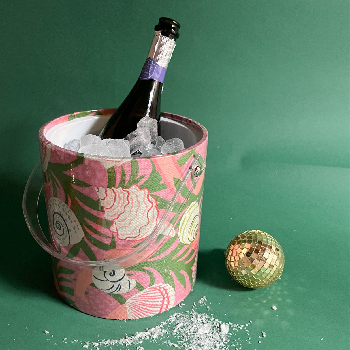 A Shell Seeker ice bucket covered in a pink and green fabric with shell motif and filled with ice and a champagne bottle