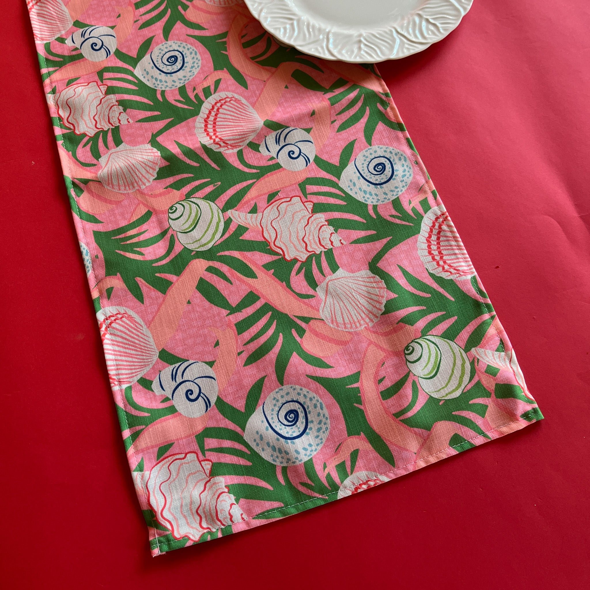 Shell Seekers cloth Table Runner features colorful shells, pink ribbons and green foliage on a pink background