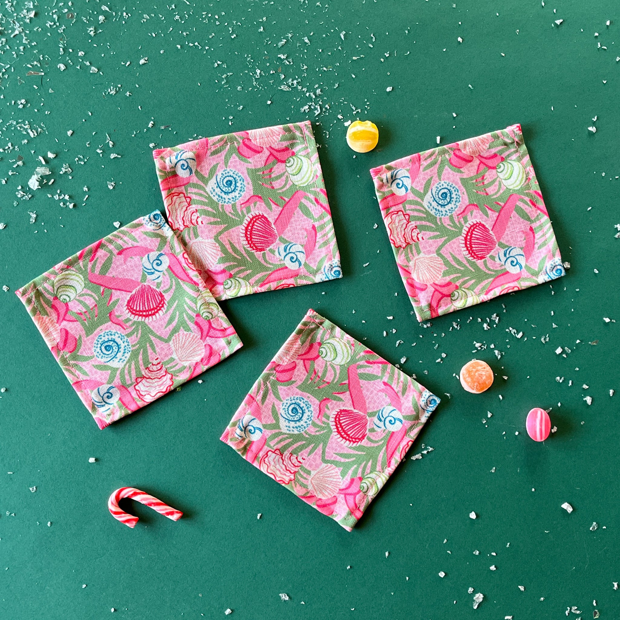 A set of four pink Shell Seekers cloth cocktail napkins with a floral and ribbon pattern, displayed on a green surface.