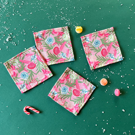 A set of four pink Shell Seekers cloth cocktail napkins with a floral and ribbon pattern, displayed on a green surface.