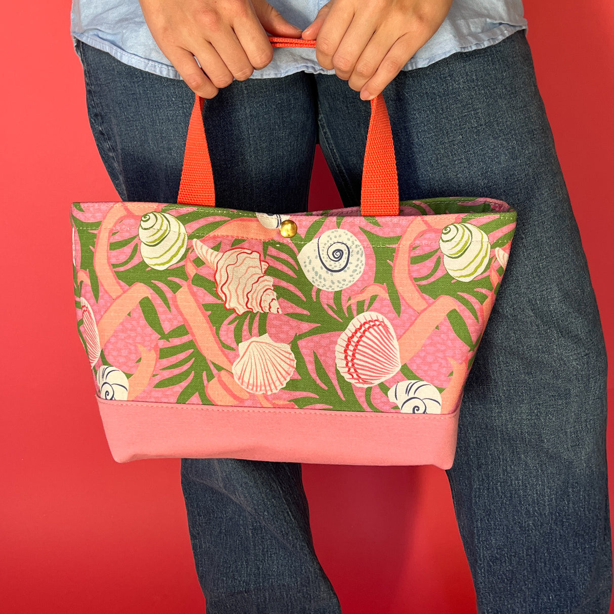 Model holding Shell Seekers canvas tote bag with pink fabric with a repeating seaweed and shell motif at leg height.