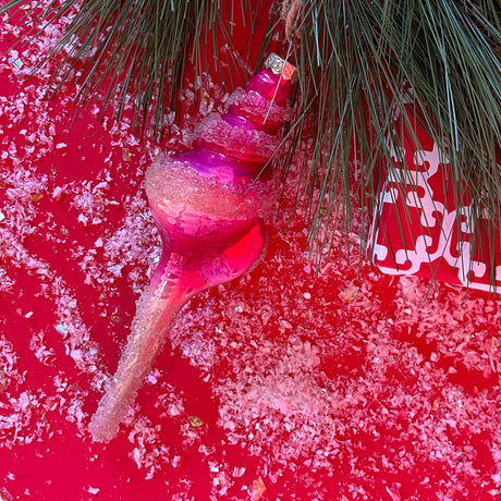 Shell-shaped Pink Glass Christmas Ornament on a red backdrop with snow and pine leaves