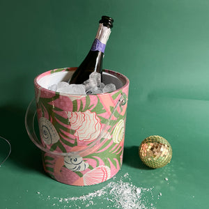 An ice bucket covered in a pink and green fabric with shell motif and filled with ice and a champagne bottle