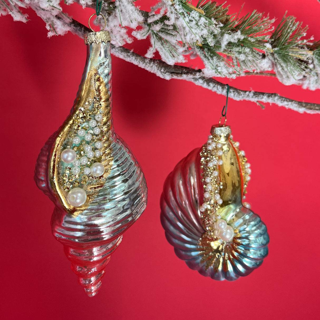 Two Shell shaped Glass Christmas Tree Ornaments with pearl embellishments hanging from a snow covered pine branch against a red background