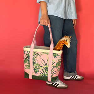 Side view of a model holding the Jungle Road large canvas tote in pink and green with a toy giraffe sticking out of the tote.