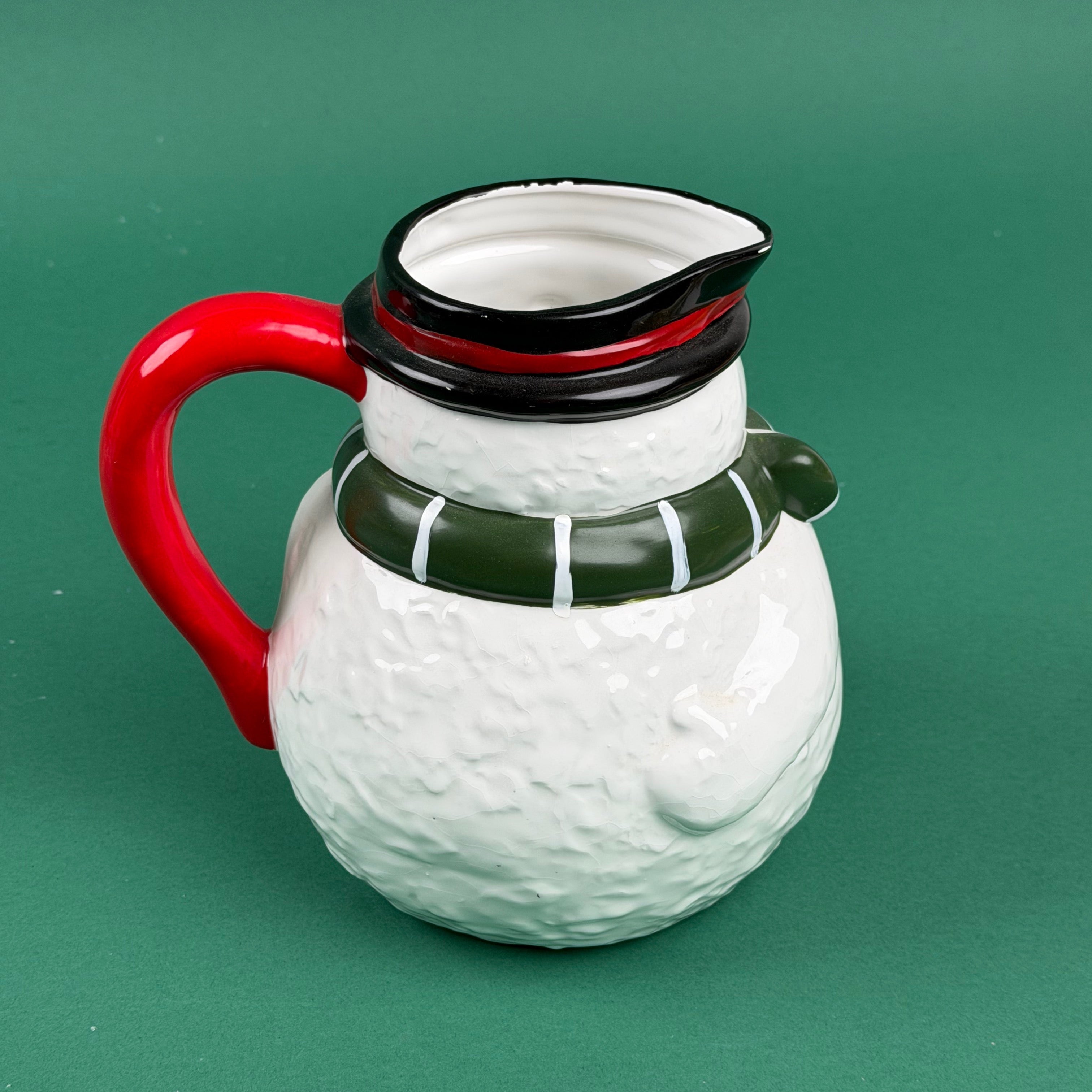 Side view of a ceramic snowman-shaped pitcher with a red handle, green scarf,on a green background