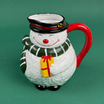 Close-up of a ceramic snowman-shaped pitcher with a red handle, green scarf, and yellow gift detail on a green background