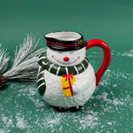 Ceramic snowman-shaped pitcher with a red handle and green scarf on a green background with faux snow and pine accents