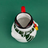 Top Close-up of a ceramic snowman-shaped pitcher with a red handle, green scarf, and yellow gift detail on a green background
