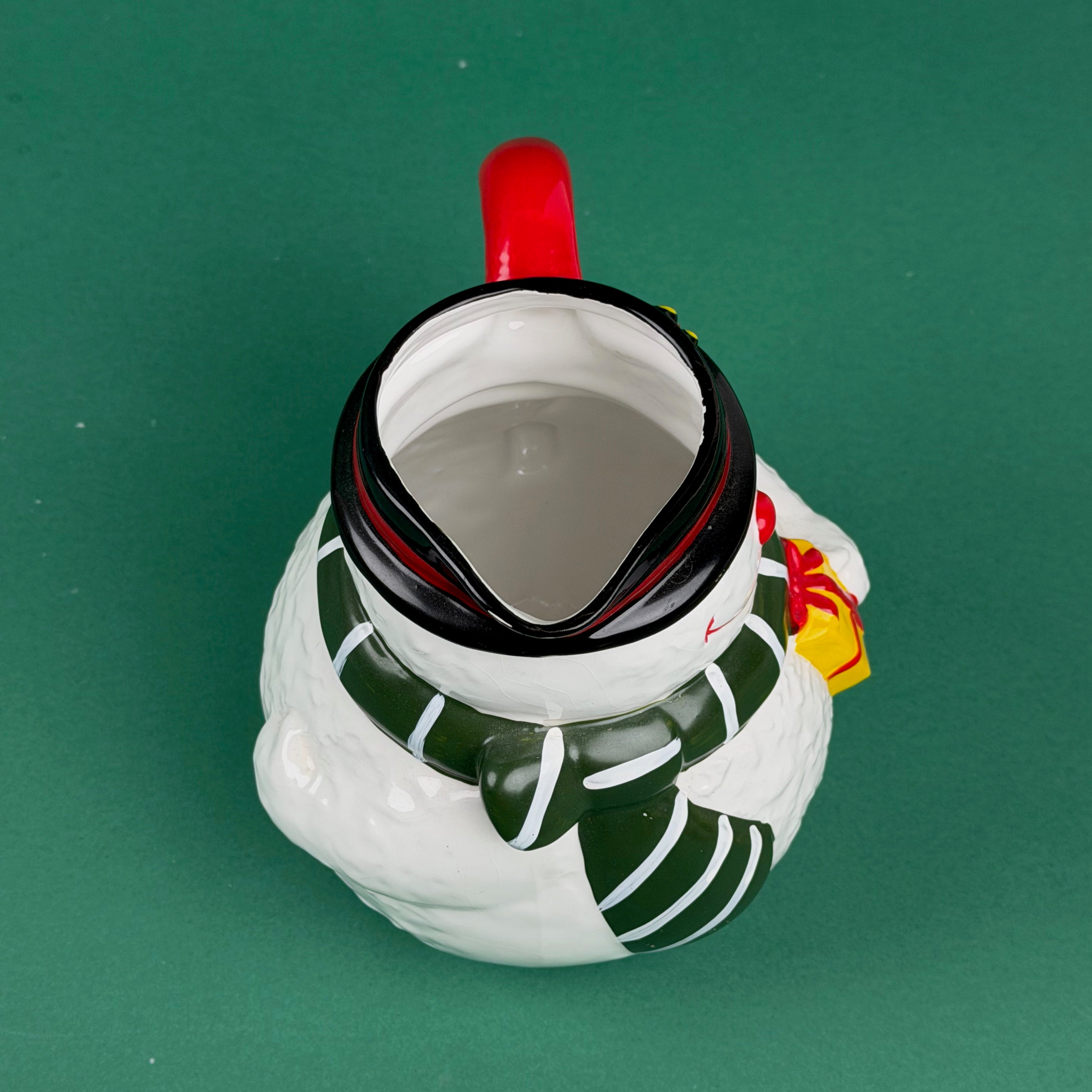 Top Close-up of a ceramic snowman-shaped pitcher with a red handle, green scarf, and yellow gift detail on a green background