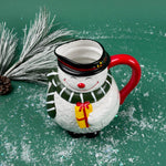 Ceramic snowman-shaped pitcher with a red handle and green scarf on a green background with faux snow and pine accents