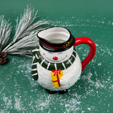 Ceramic snowman-shaped pitcher with a red handle and green scarf on a green background with faux snow and pine accents